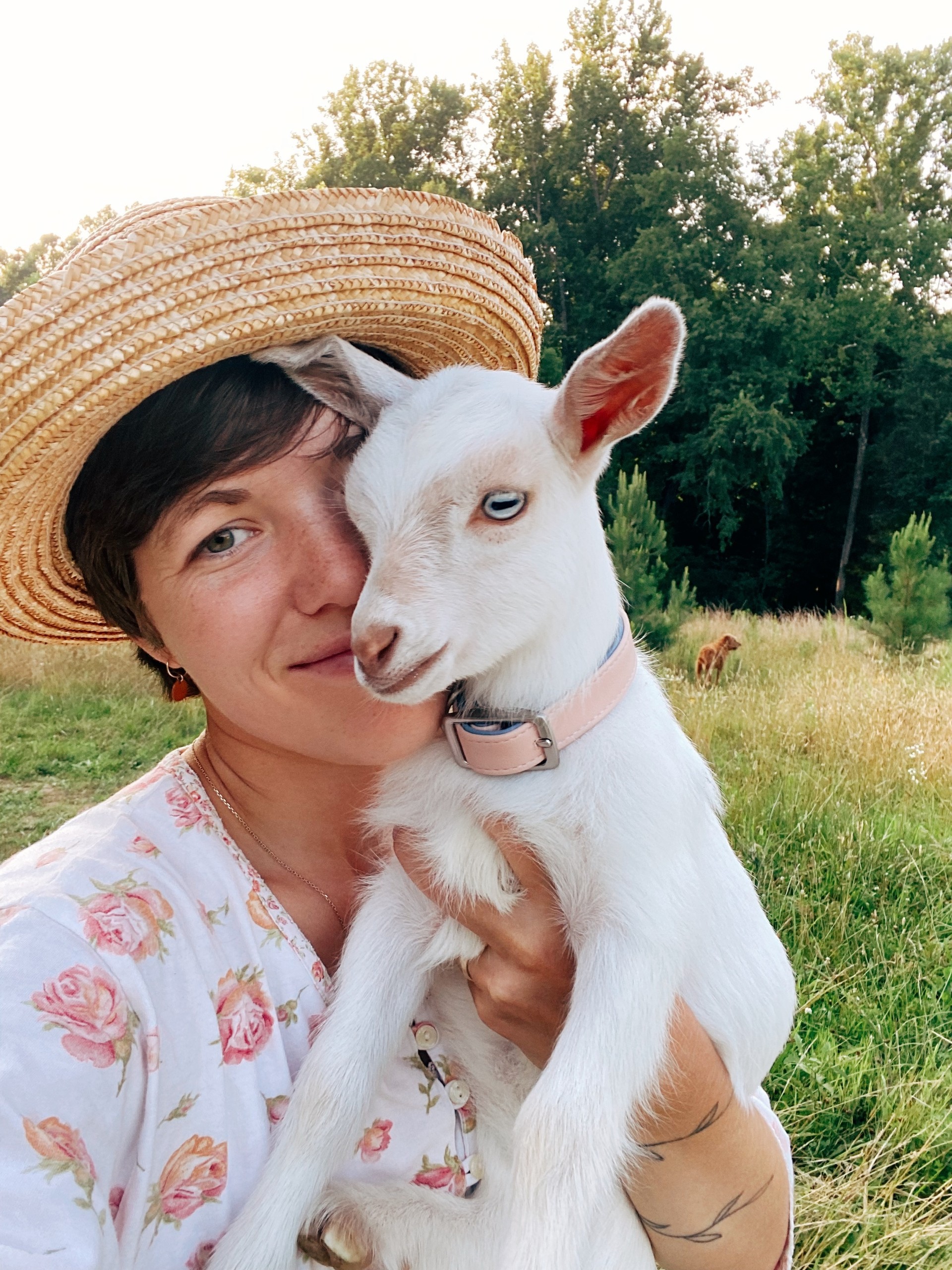 Person in straw hat holds a baby goat in a grassy field.