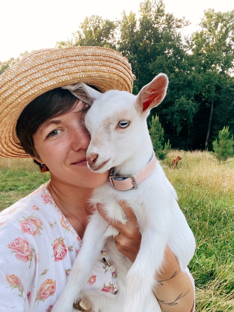Person in straw hat holds a baby goat in a grassy field.